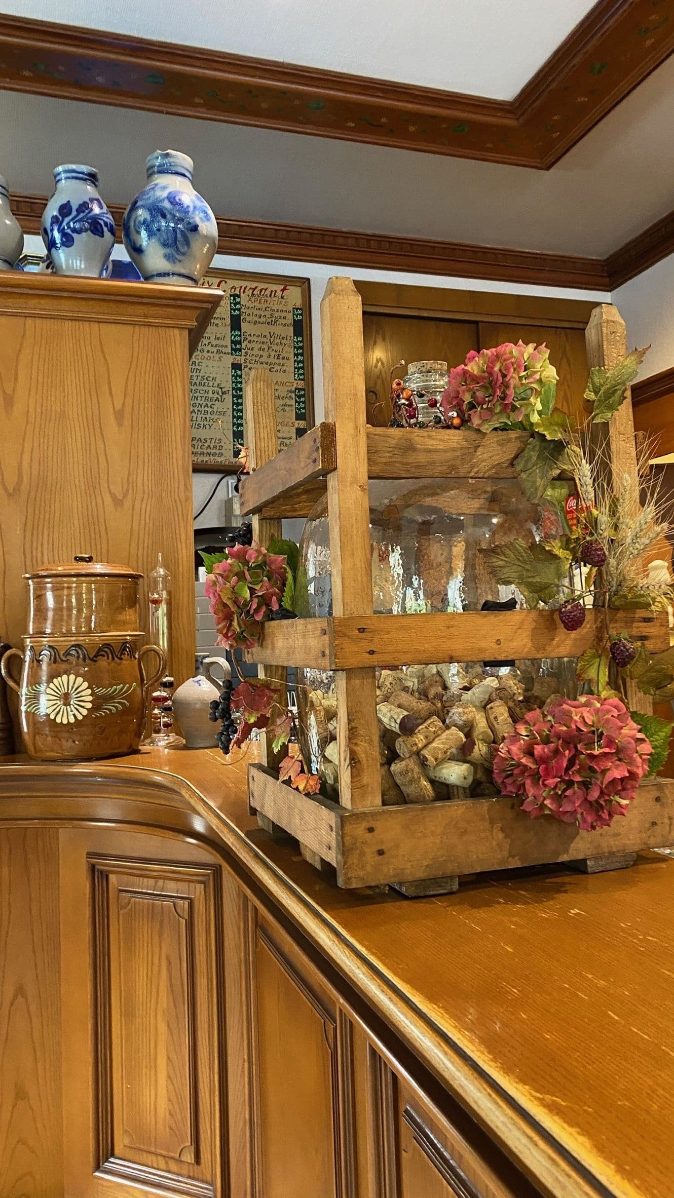Vitrine chaleureuse du comptoir avec un grand bocal en verre rempli de bouchons de vin dans une caisse en bois, décoré d'hortensias séchés et de poteries alsaciennes sur un comptoir en bois verni.