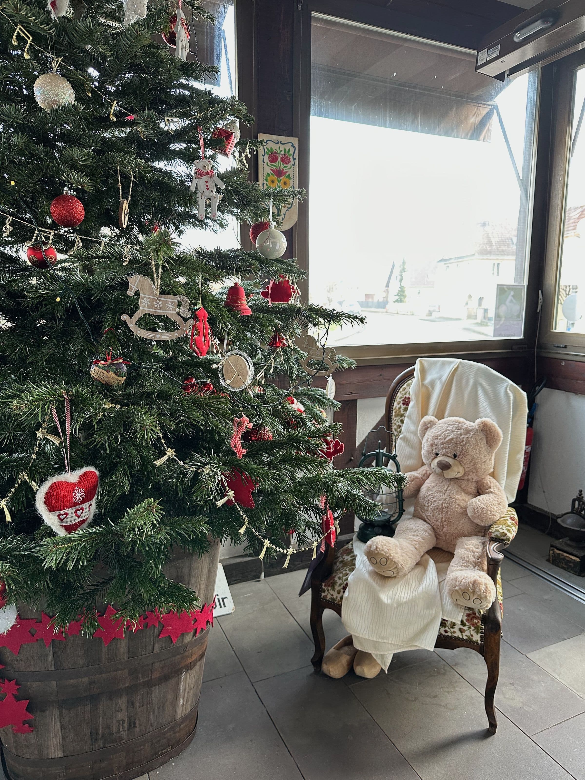 Coin chaleureux et festif de l'hôtel avec un grand sapin de Noël décoré dans un baril en bois, à côté d'un fauteuil ancien portant un gros ours en peluche et un plaid blanc.