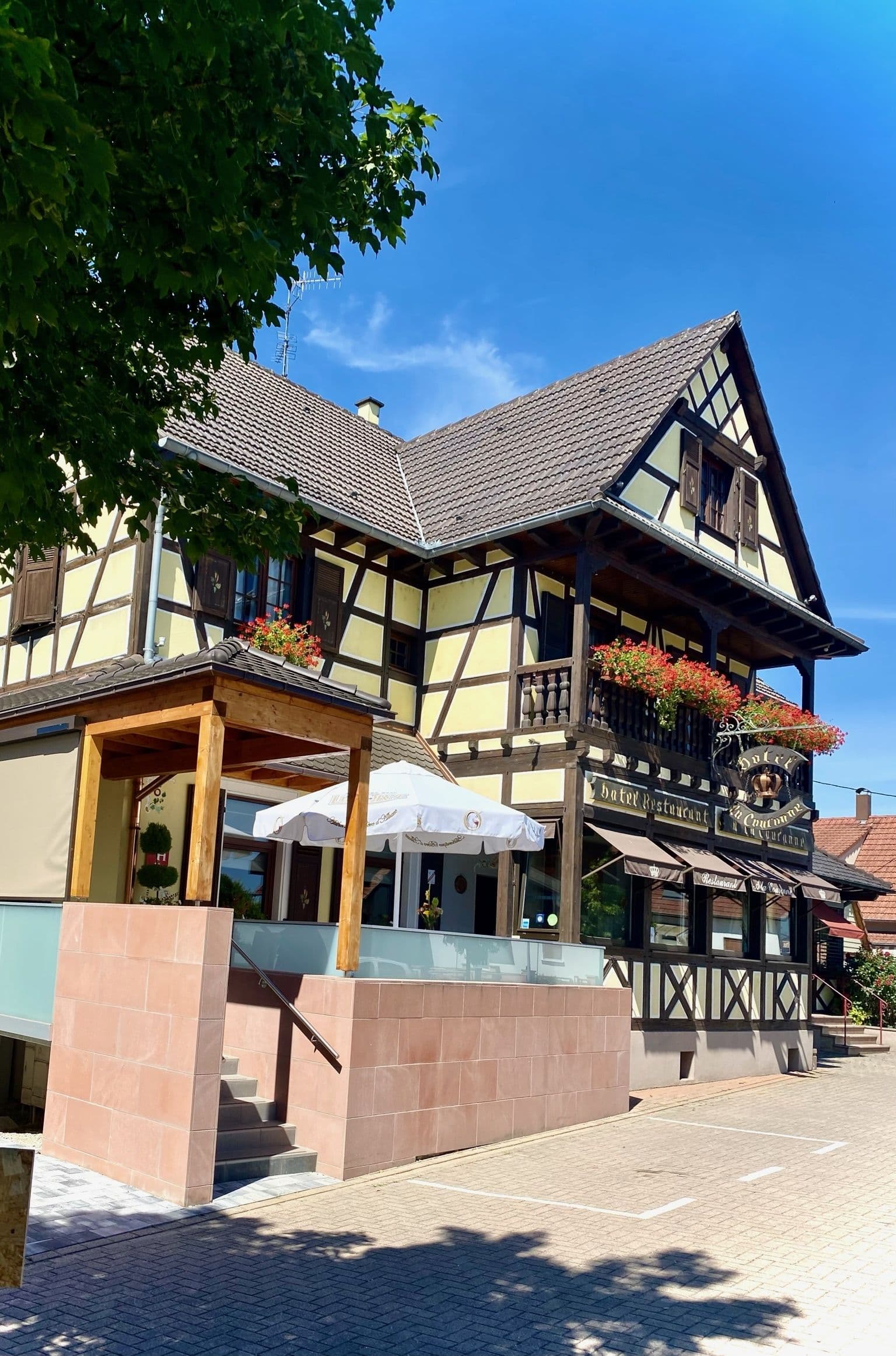 Façade de l'hôtel-restaurant alsacien à colombages par une journée ensoleillée, balcon fleuri et terrasse extérieure avec parasol devant l'entrée.