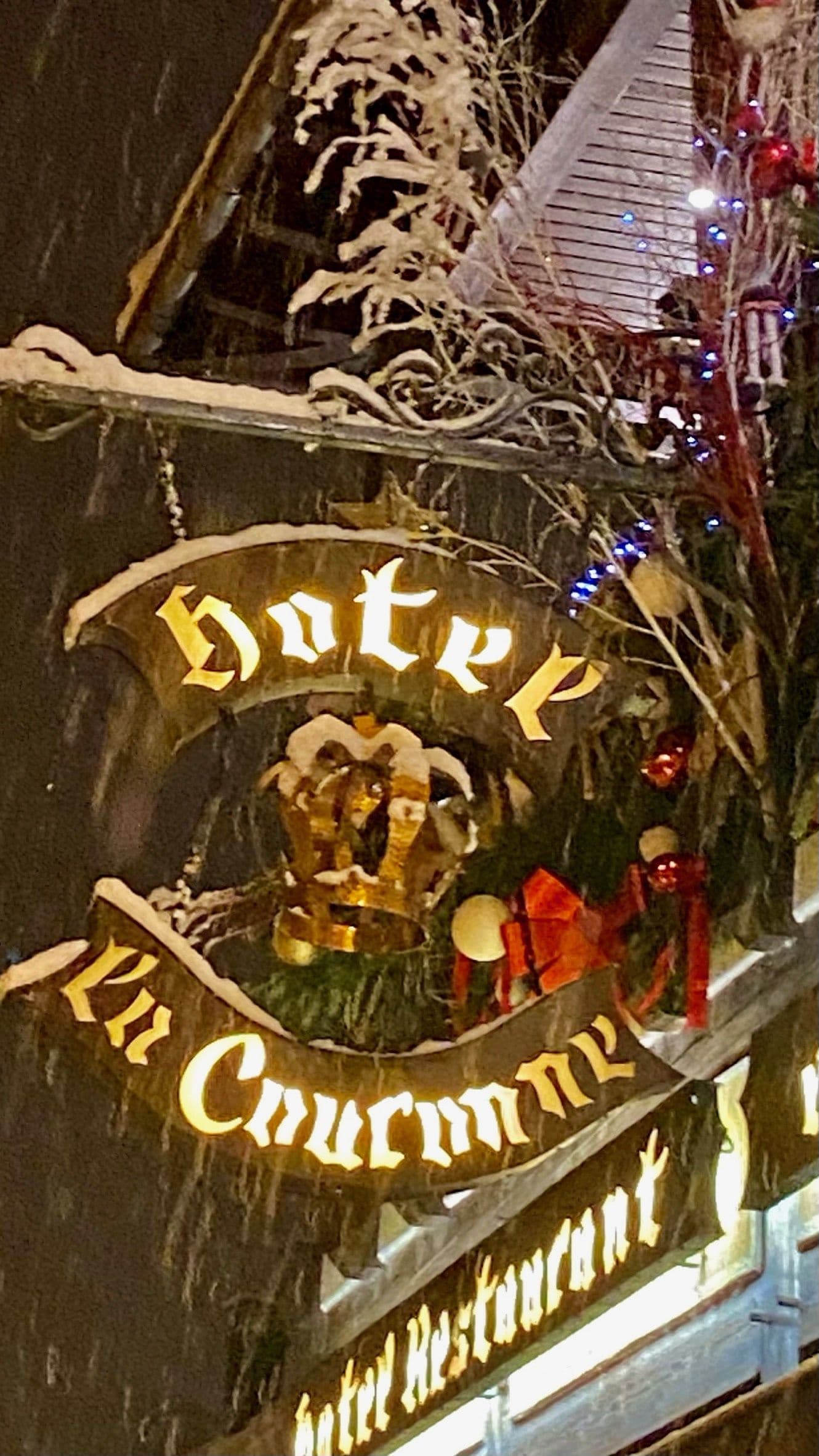 Gros plan nocturne de l'enseigne lumineuse de l'hôtel « À La Couronne », ornée d'une couronne et saupoudrée de neige, entourée de guirlandes et décorations de saison.
