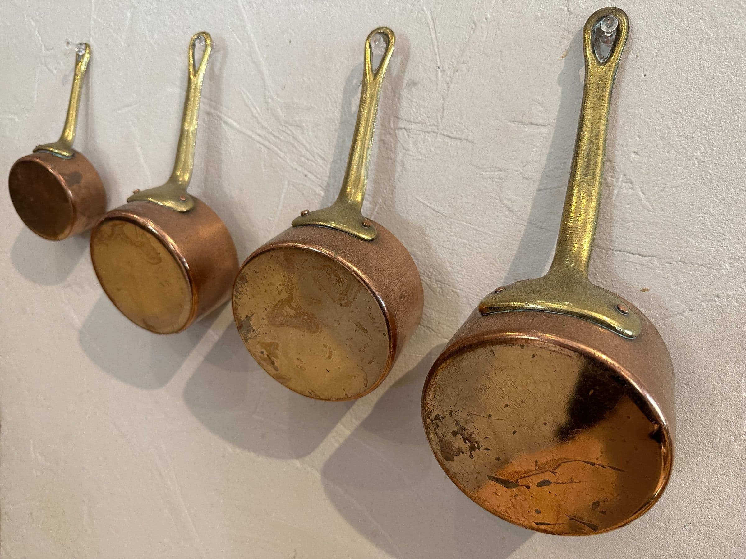 Quatre casseroles en cuivre anciennes aux manches en laiton accrochées sur un mur en plâtre texturé, détail chaleureux et rustique de la cuisine traditionnelle.