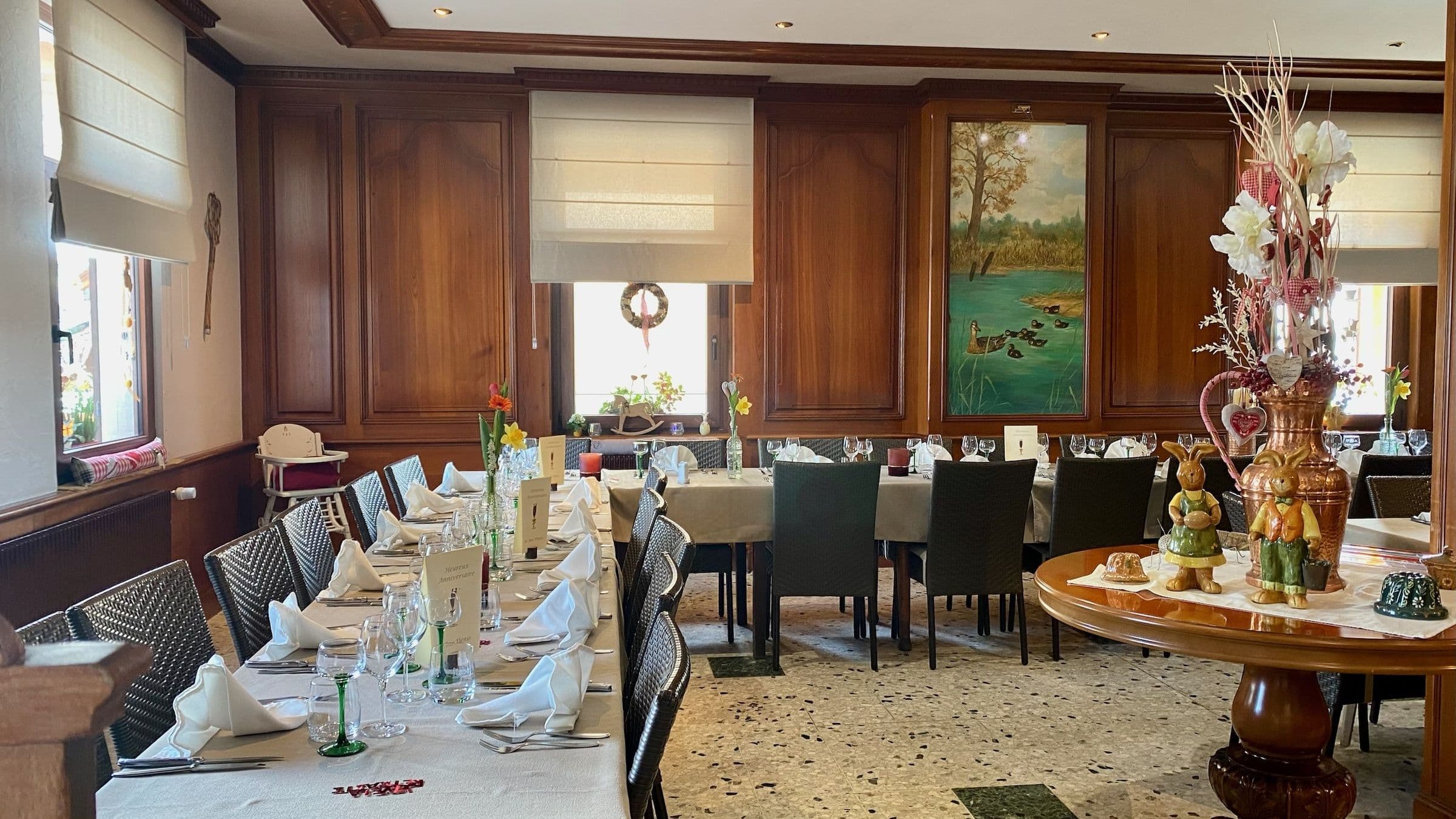 Salle à manger alsacienne chaleureuse avec longues tables dressées, nappes blanches, verres et vases fleuris, boiseries et décorations printanières.