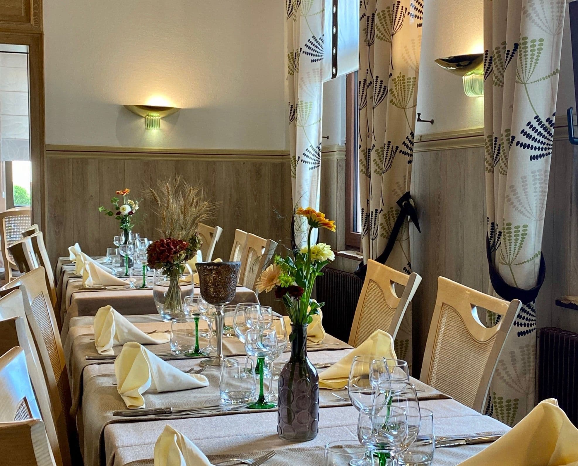 Table de restaurant élégamment dressée avec serviettes pliées, verres et petites compositions florales, éclairage doux et rideaux à motifs près d'une fenêtre.