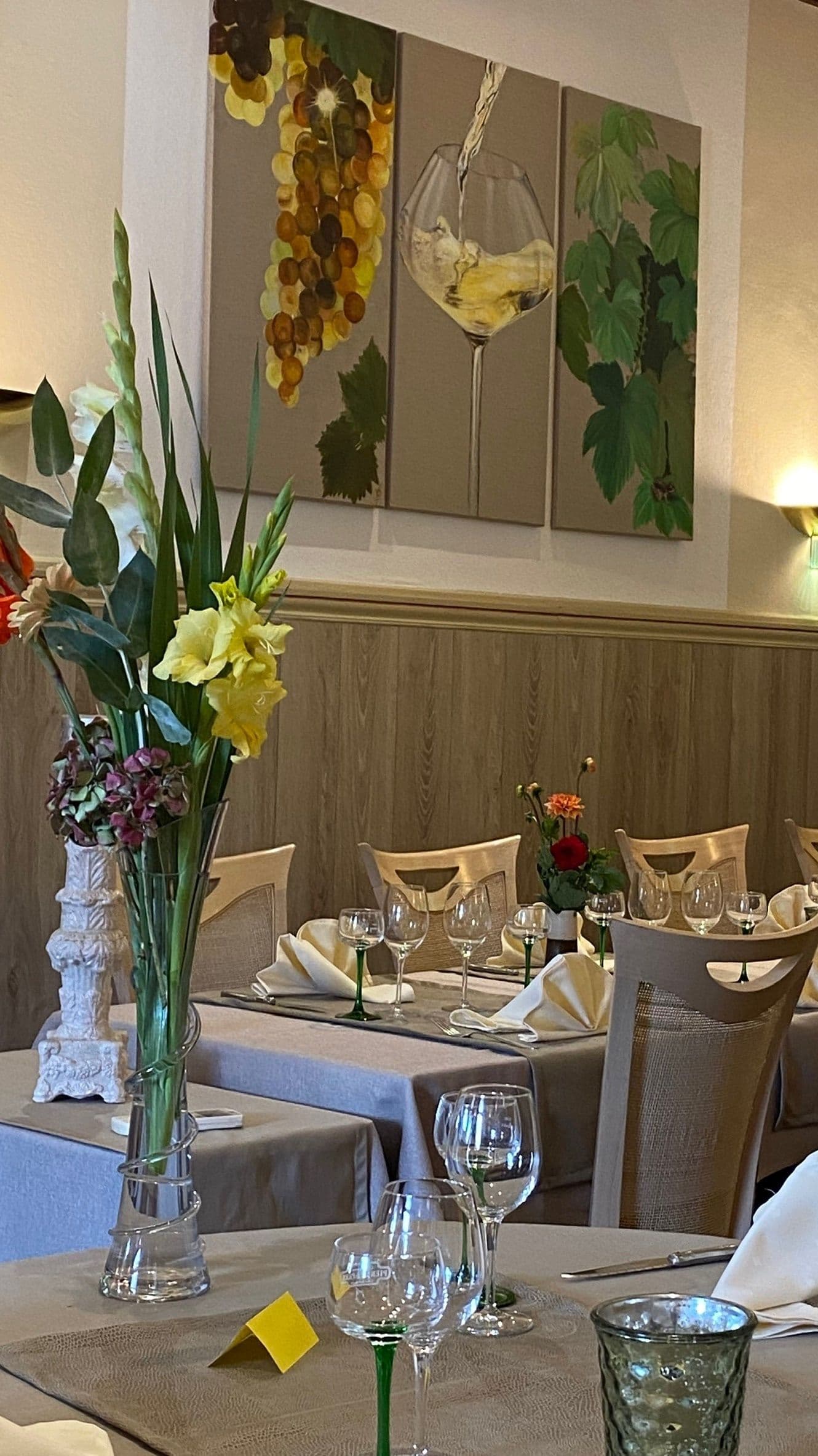 Salle à manger chaleureuse dressée pour le service, tables avec verres à vin aux pieds verts, nappes et bouquets de fleurs sous un triptyque représentant des grappes et un verre de vin.