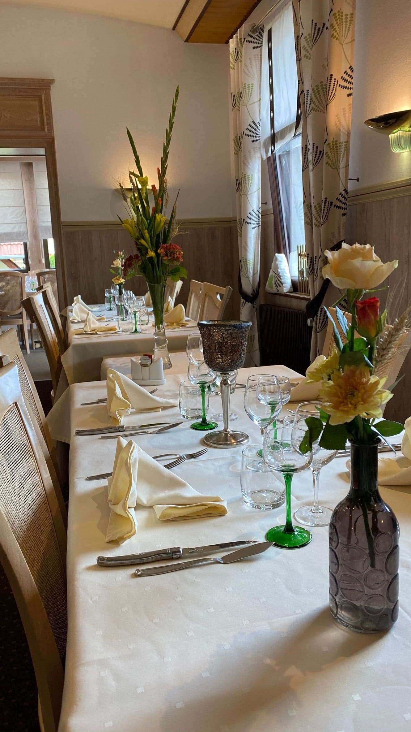 Salle à manger chaleureuse d’un hôtel-restaurant alsacien avec une table dressée en nappes blanches, serviettes pliées, verres à pied aux tiges vertes et bouquets de fleurs, prête à recevoir des convives.
