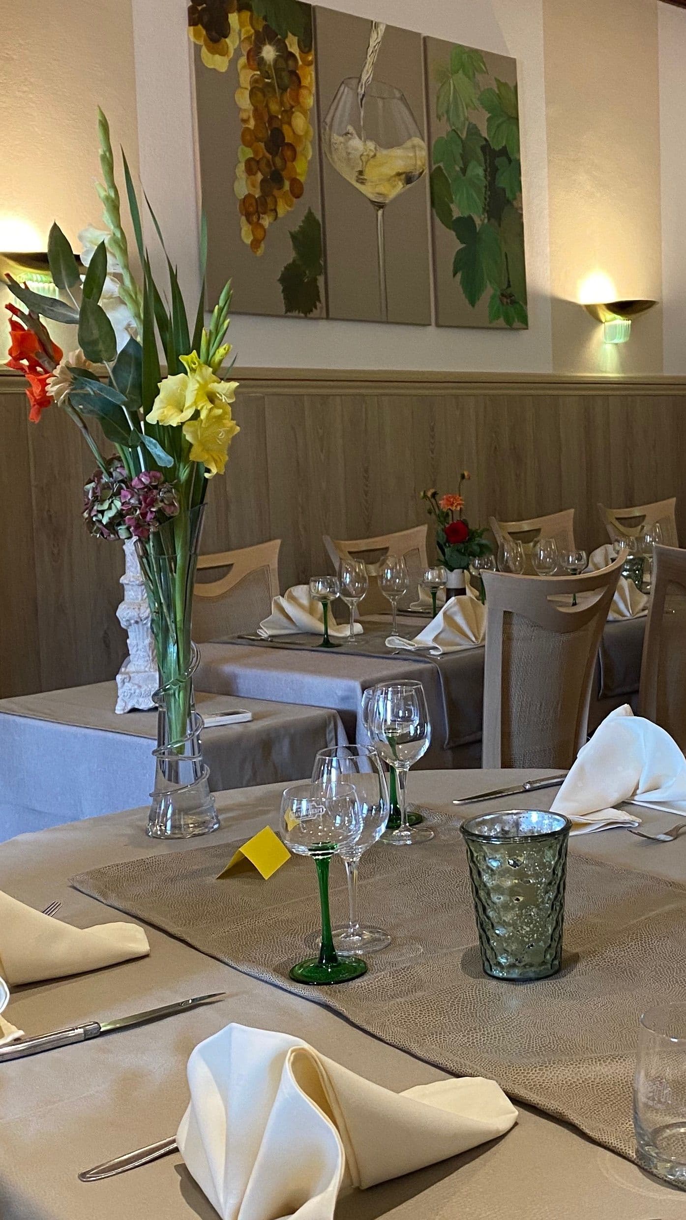 Salle à manger traditionnelle d'hôtel-restaurant avec tables dressées, verres à vin à tige verte, serviettes pliées et grand vase de fleurs sous un tableau représentant du raisin et du vin.