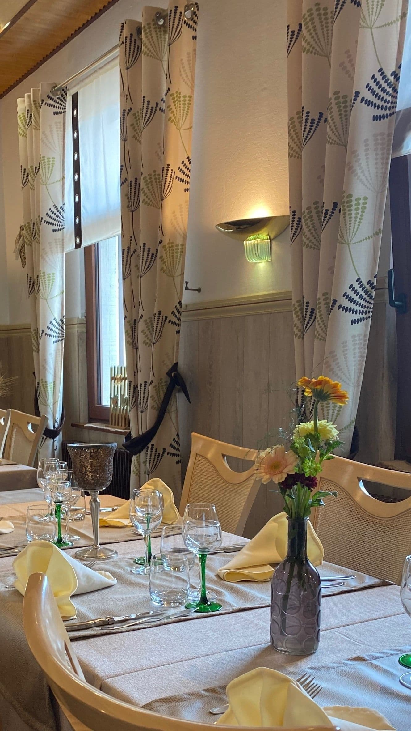 Table de restaurant dressée avec serviettes jaunes, verres à pied au bord vert et un petit vase de fleurs devant des rideaux à motifs et une fenêtre en bois, éclairée par une applique murale chaude.