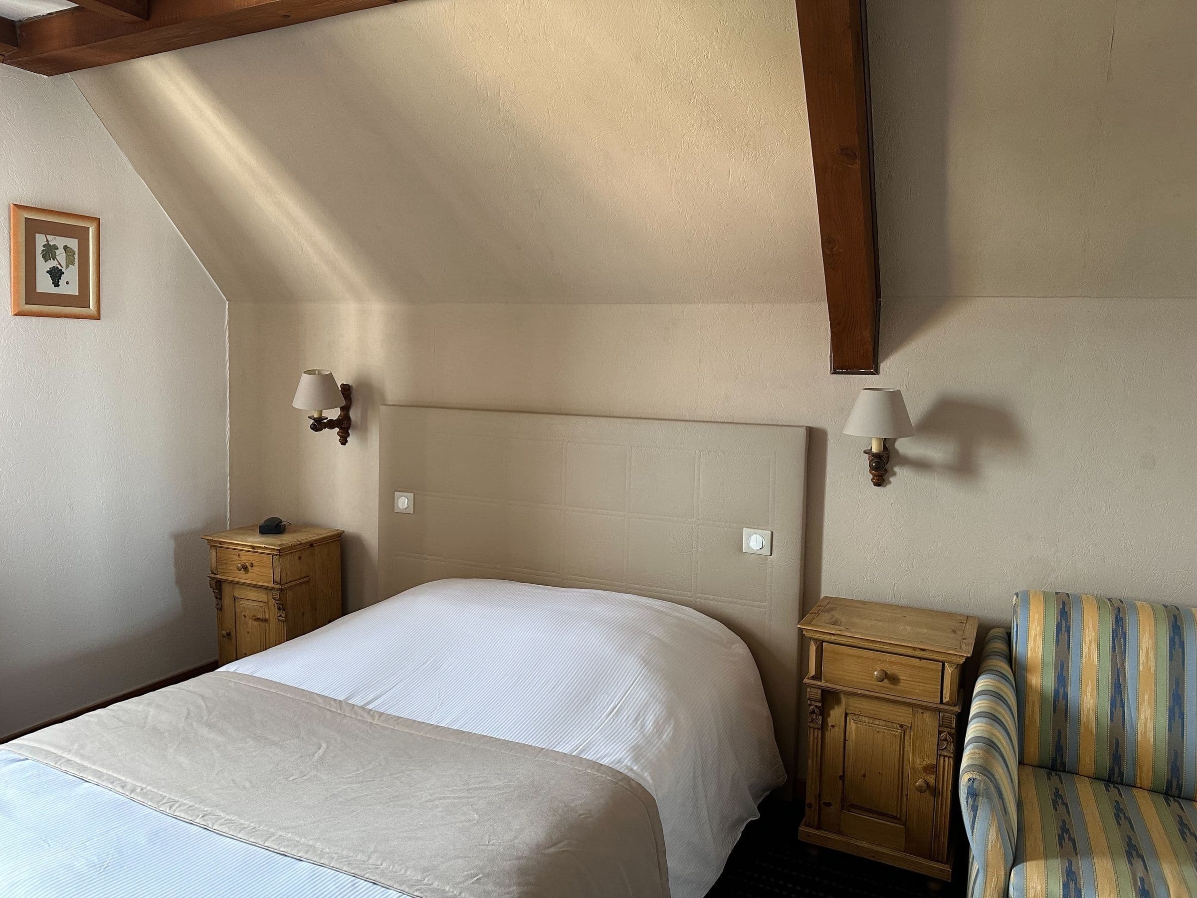 Chambre double sous combles avec poutres apparentes, tête de lit rembourrée, deux tables de chevet en bois rustique, lampes murales et un fauteuil rayé, décor sobre et chaleureux.