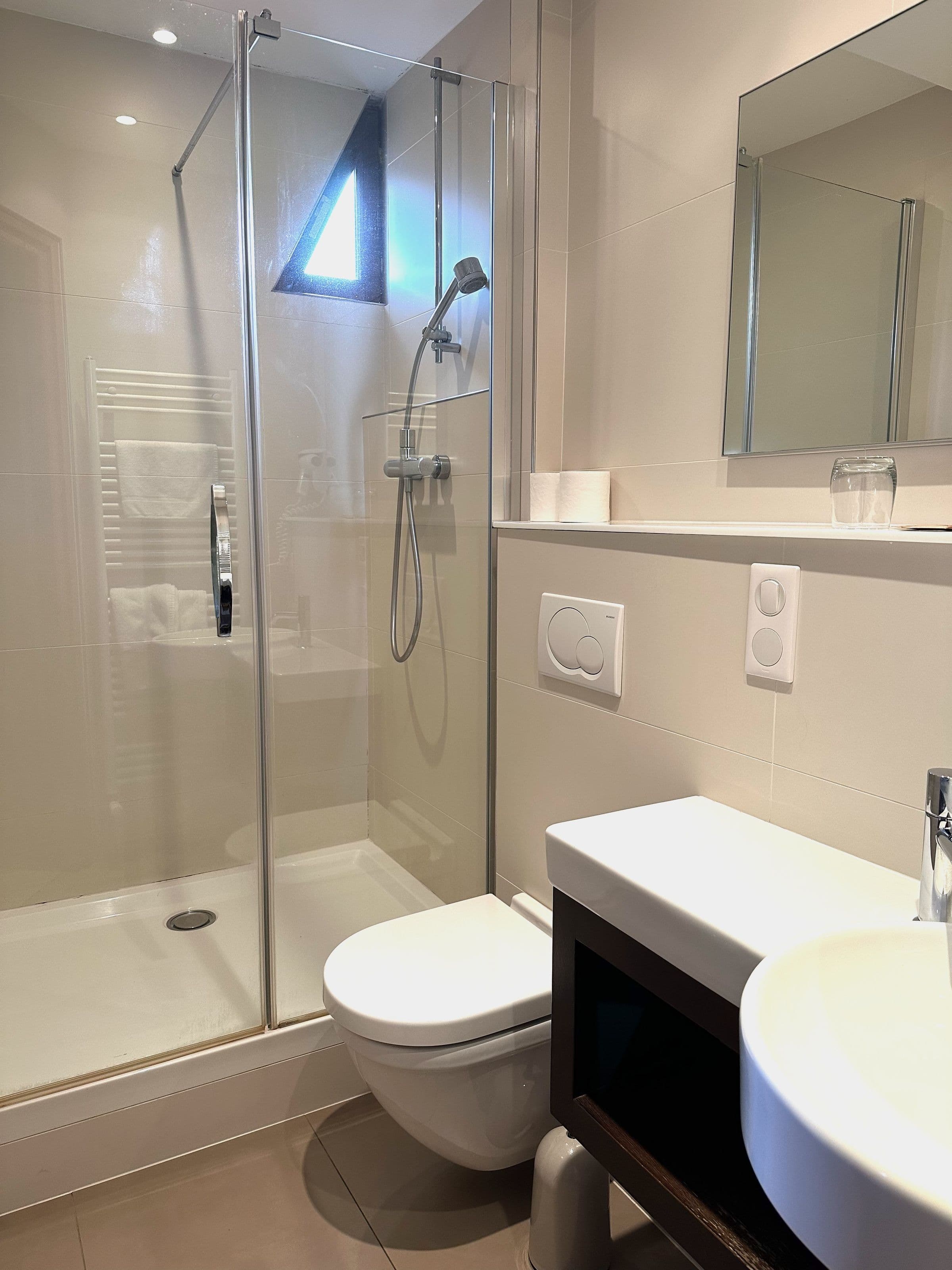Salle de bain attenante moderne avec cabine de douche vitrée, douchette, toilette suspendue et petite fenêtre triangulaire apportant de la lumière naturelle.