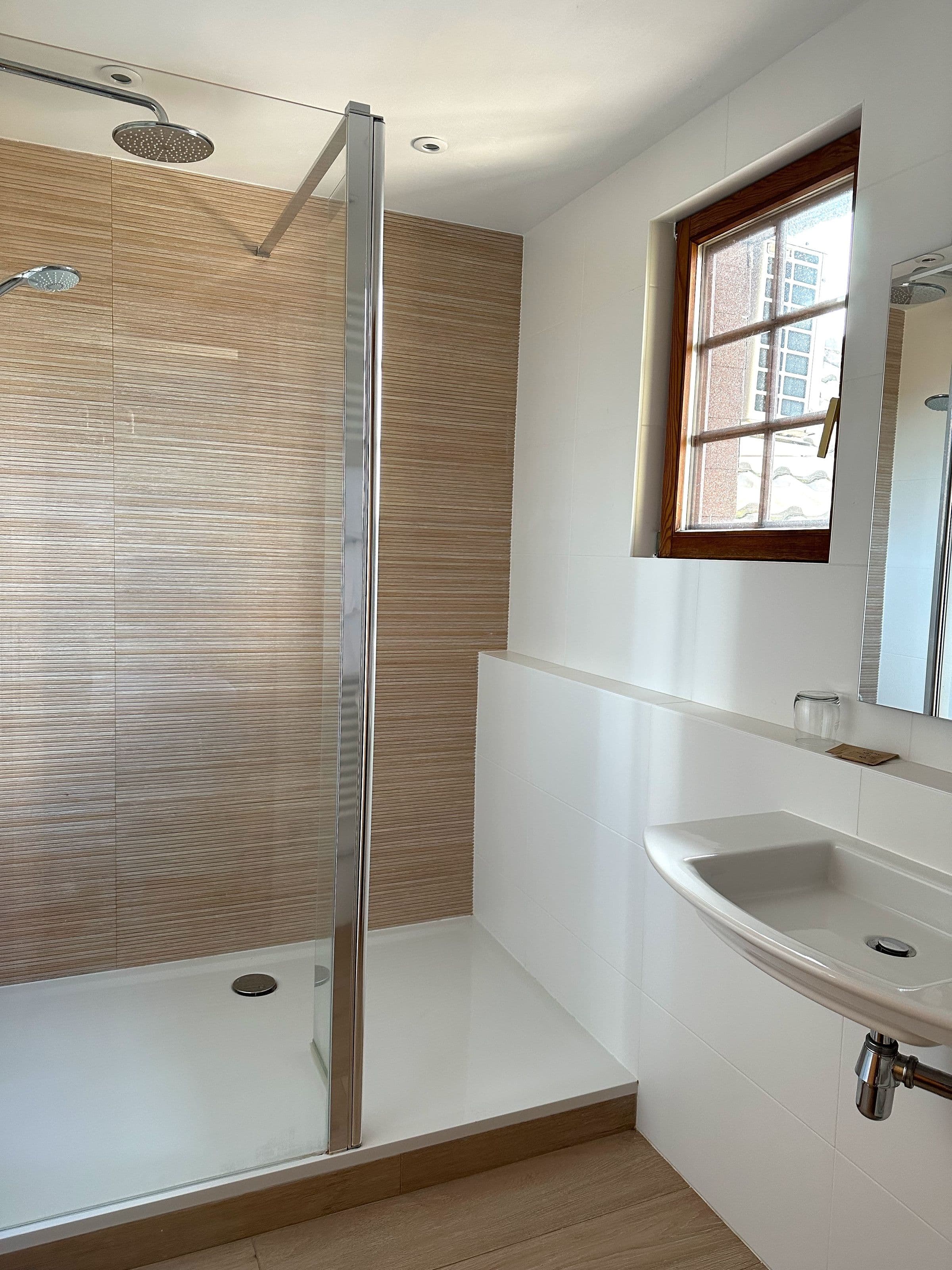 Salle de bain lumineuse avec douche à l'italienne, pomme de douche pluie, paroi vitrée et carrelage imitation bois, lavabo mural et fenêtre à cadre bois apportant de la lumière naturelle.