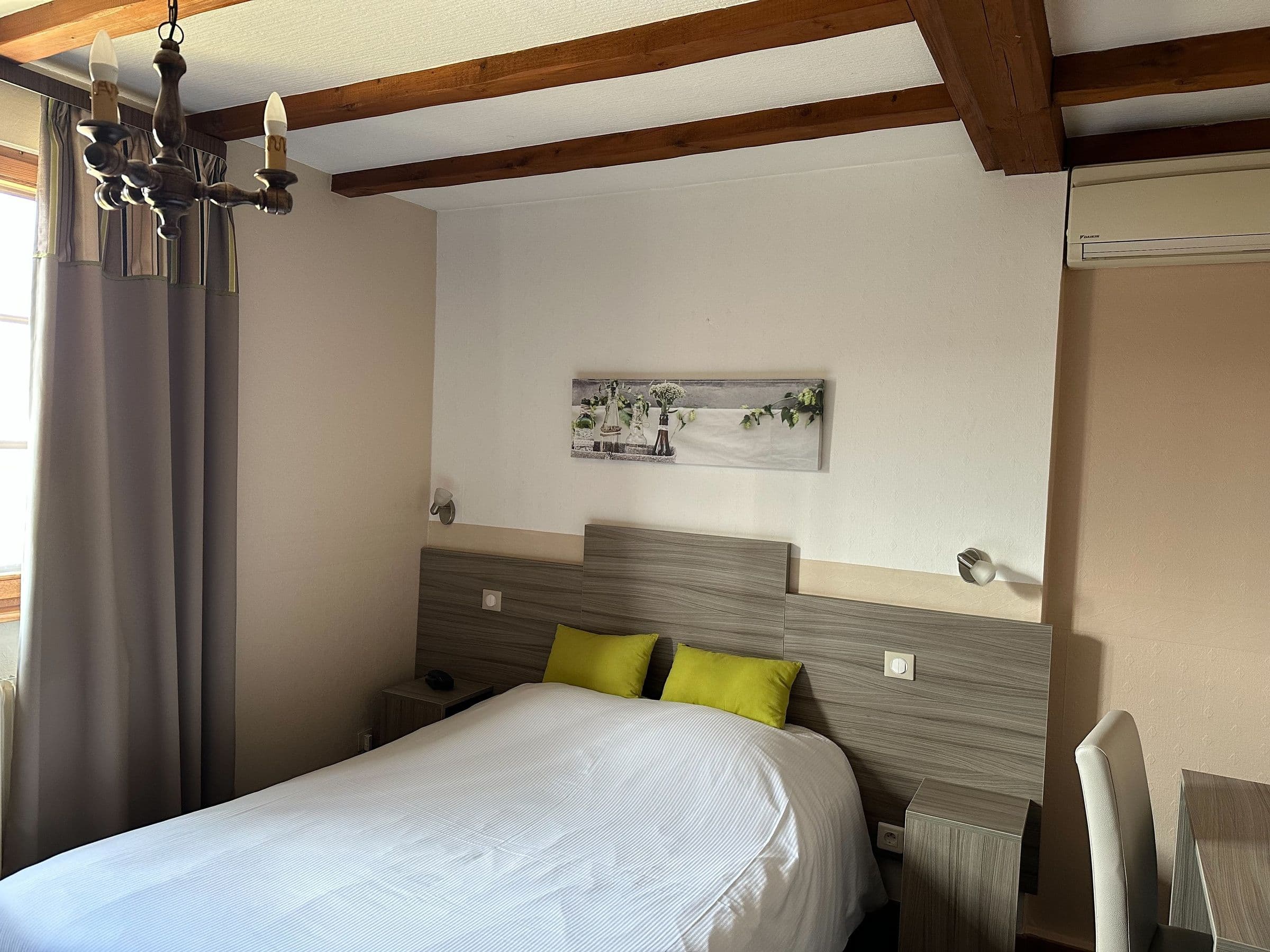 Chambre double chaleureuse avec poutres apparentes, tête de lit imitation bois, deux coussins vert anis et une fenêtre à rideaux laissant entrer la lumière naturelle.