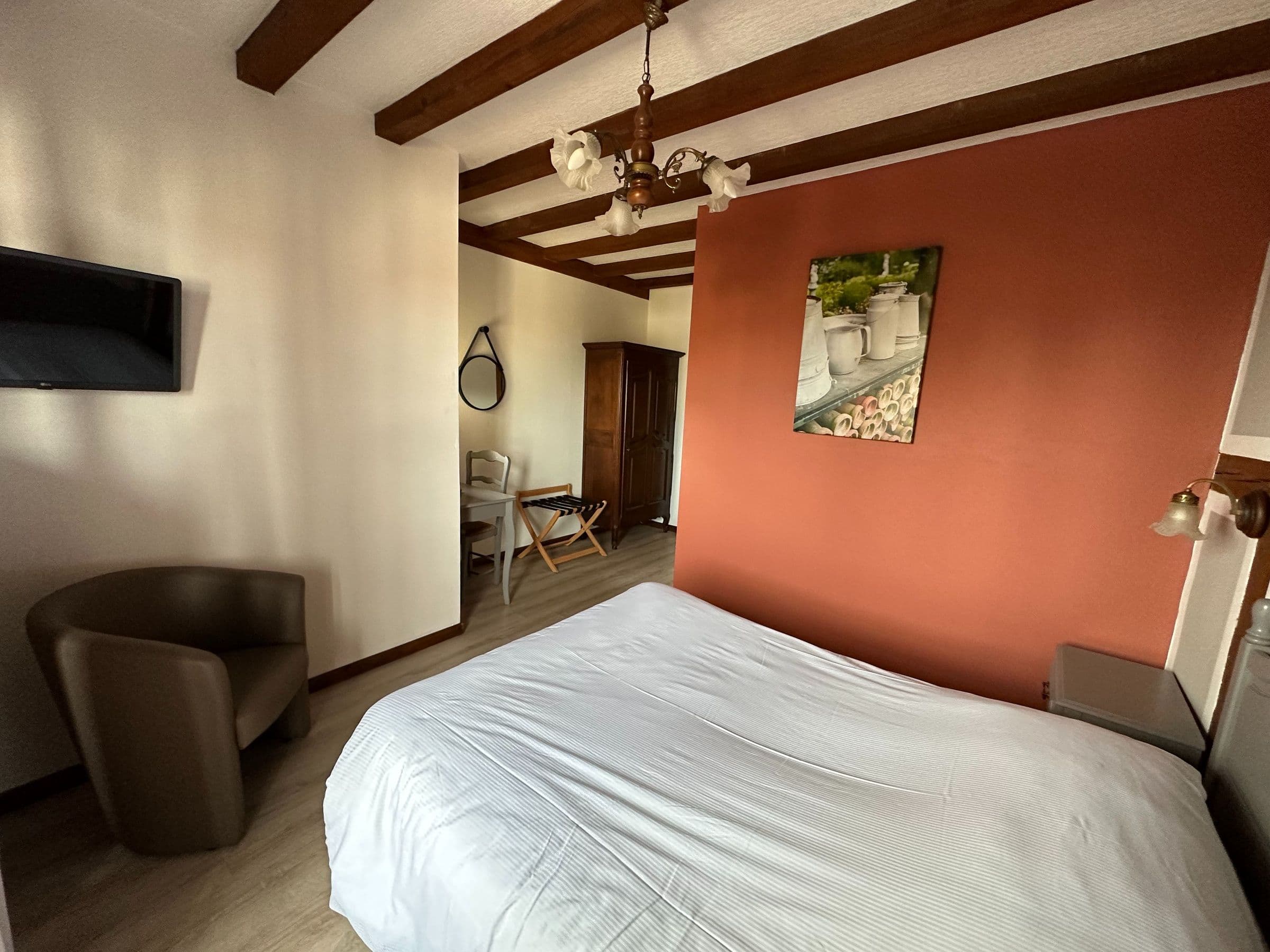 Chambre double lumineuse avec poutres apparentes, mur d’accent terracotta, lit en draps blancs, fauteuil et coin bureau, ambiance chaleureuse et traditionnelle alsacienne.