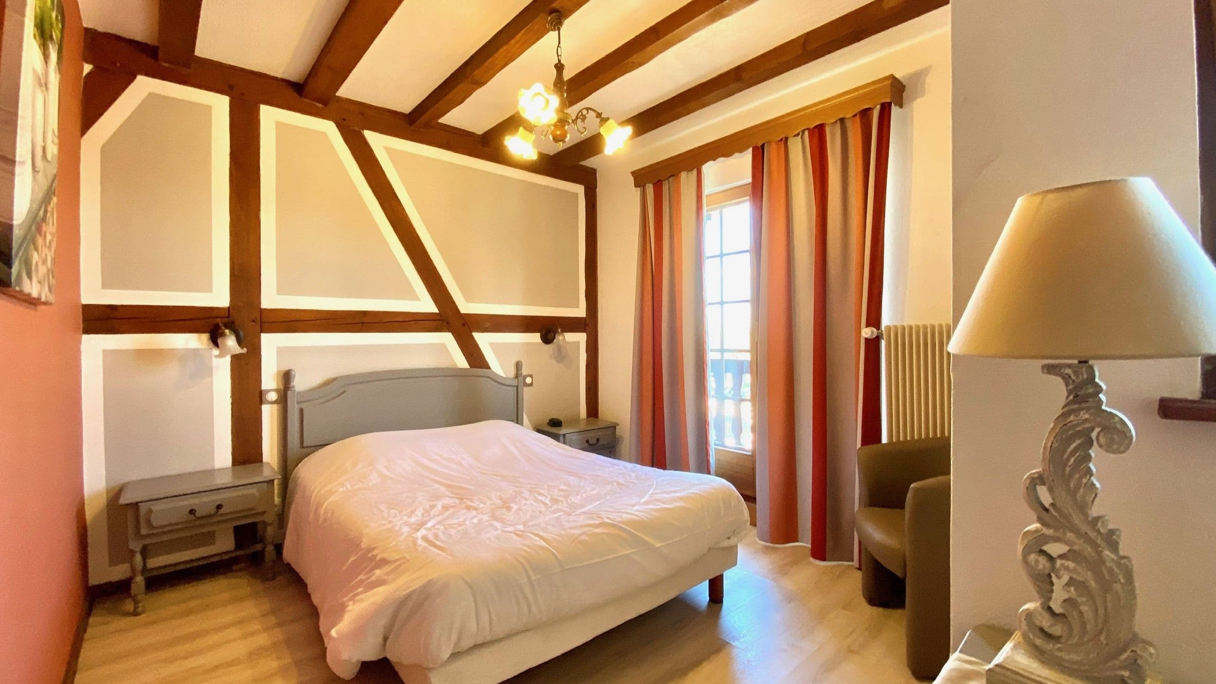 Chambre double lumineuse aux poutres apparentes et décoration alsacienne traditionnelle, avec lit double, tables de chevet, fauteuil et fenêtre aux rideaux rayés laissant entrer la lumière.