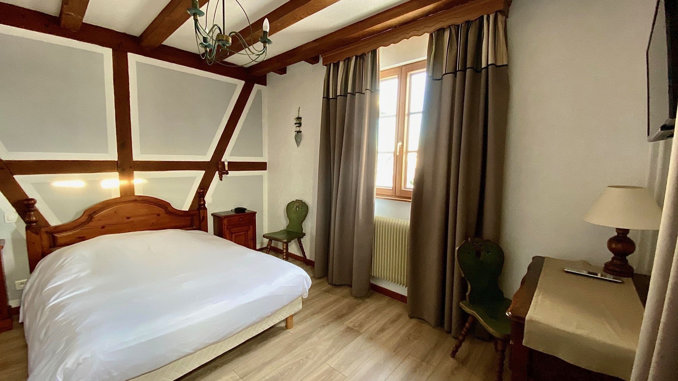 Chambre double chaleureuse avec poutres apparentes, lit bien fait et fenêtre en bois laissant entrer la lumière naturelle; mobilier traditionnel alsacien avec chaise et bureau.
