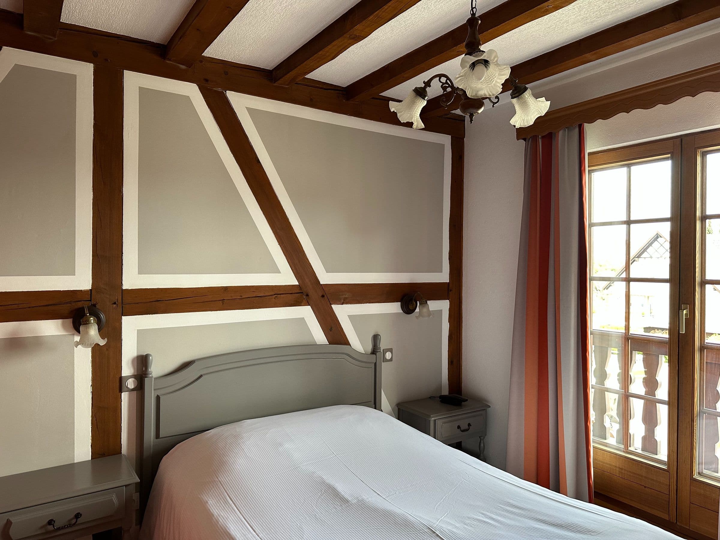 Chambre double de style traditionnel alsacien avec poutres apparentes, tête de lit grise, tables de chevet assorties et porte-fenêtre ouvrant sur un petit balcon en bois.