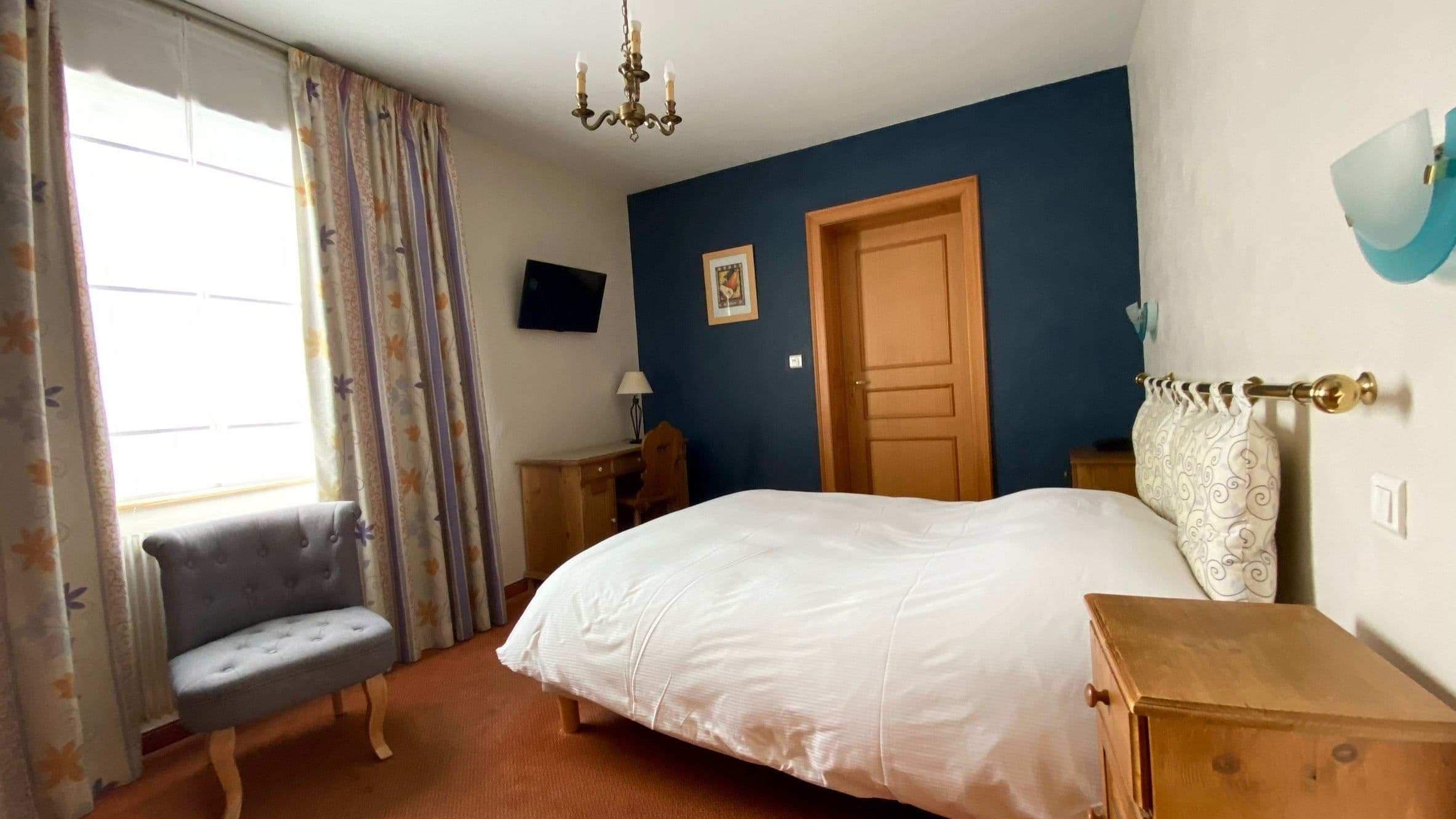 Chambre double lumineuse au style traditionnel avec lit blanc, tête de lit à motif, murs bleus et mobilier en bois, fenêtre avec rideaux floraux et fauteuil gris.