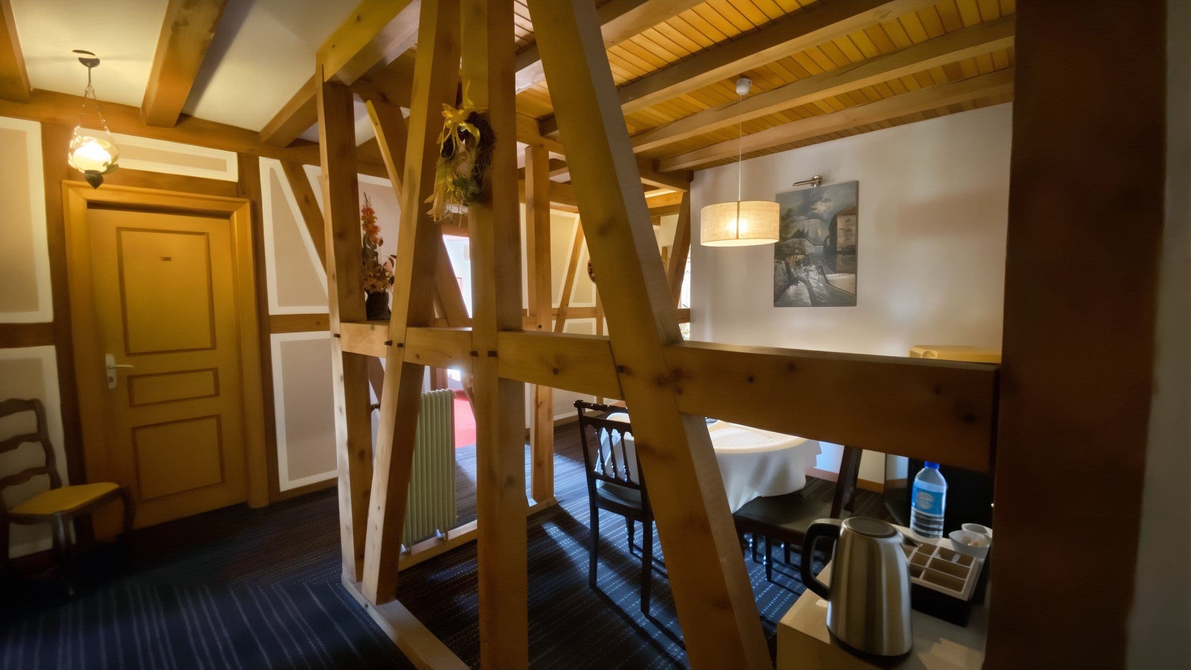 Coin repas chaleureux séparé par des poutres à colombage alsaciennes, table dressée, éclairage doux et bouilloire avec bouteille d'eau sur le meuble.