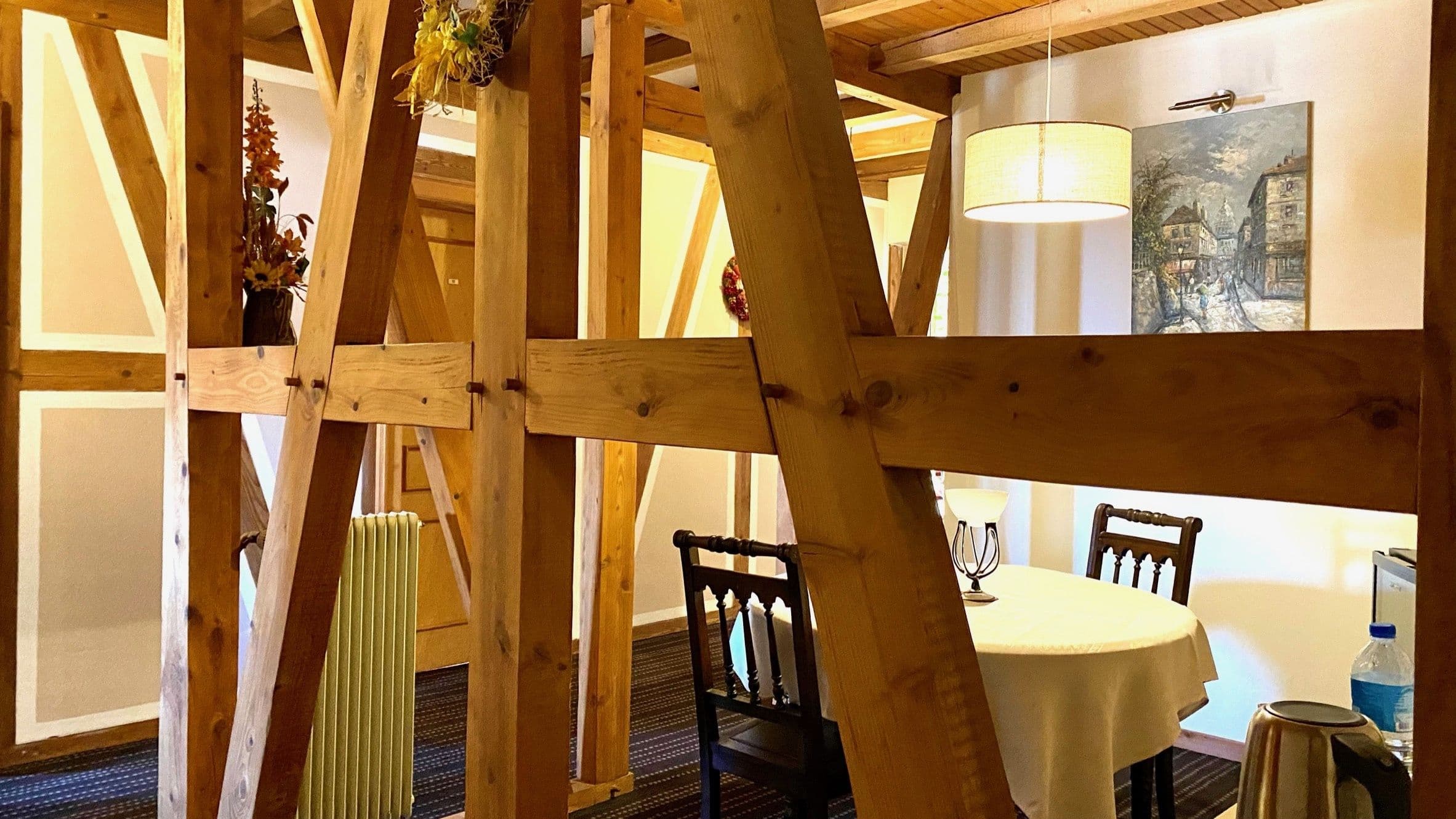 Intérieur chaleureux à colombages apparents séparant un coin repas avec une table ronde nappée et des chaises anciennes, éclairé par une lampe suspendue.