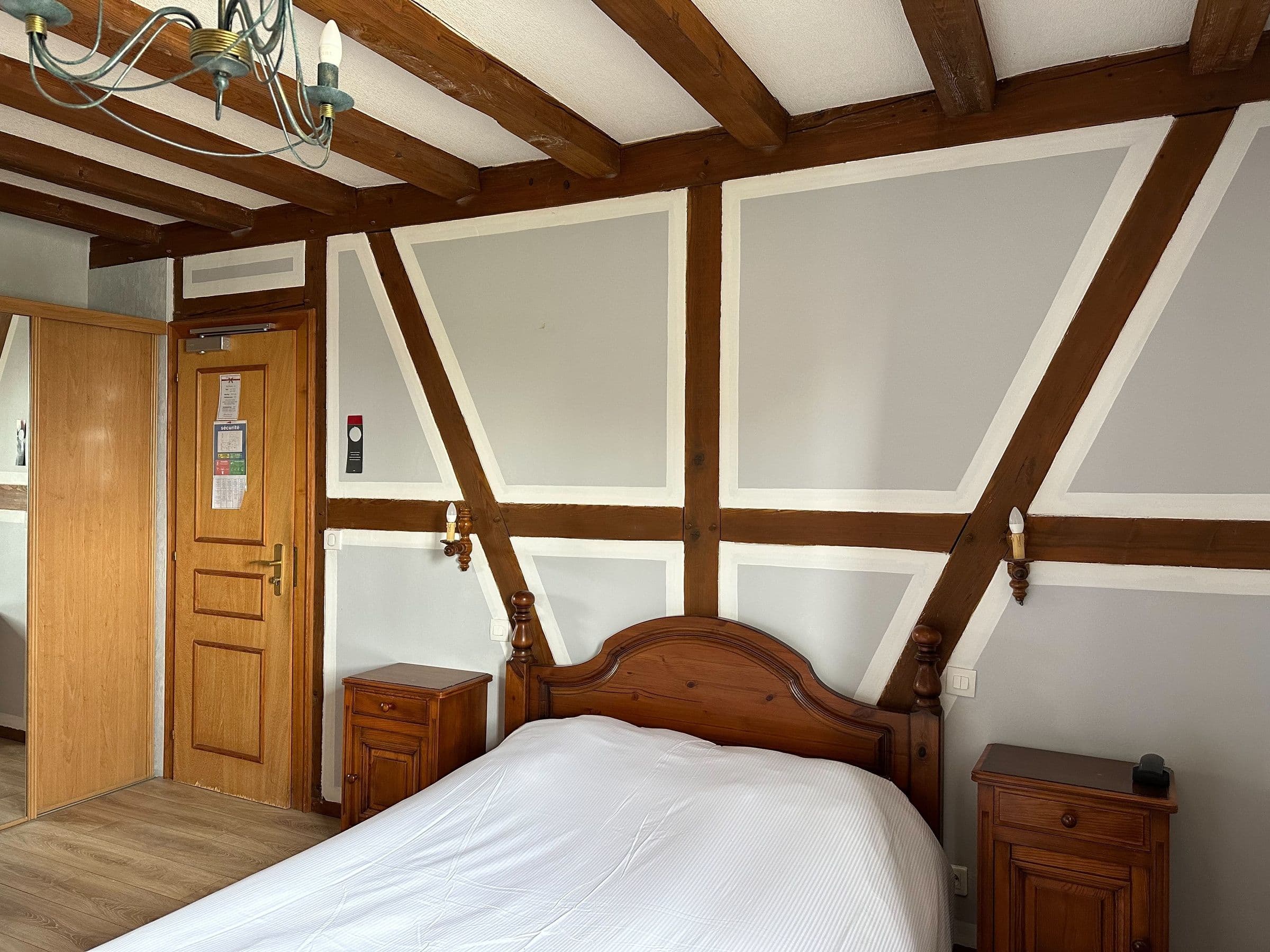 Chambre double traditionnelle avec poutres apparentes alsaciennes, tête de lit en bois, tables de chevet assorties et sol en parquet, éclairage discret et armoire près de la porte.