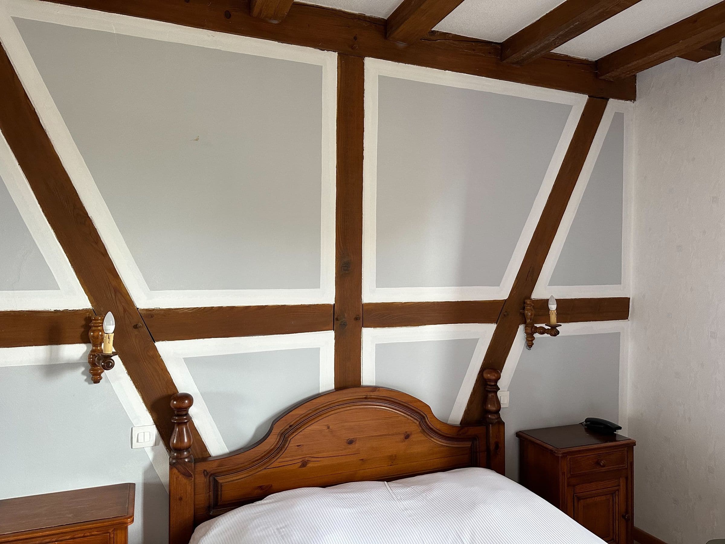 Coin de chambre d'hôtel traditionnel avec poutres apparentes foncées, panneaux clairs, tête de lit en pin sculpté et tables de chevet assorties, éclairage mural et literie blanche.