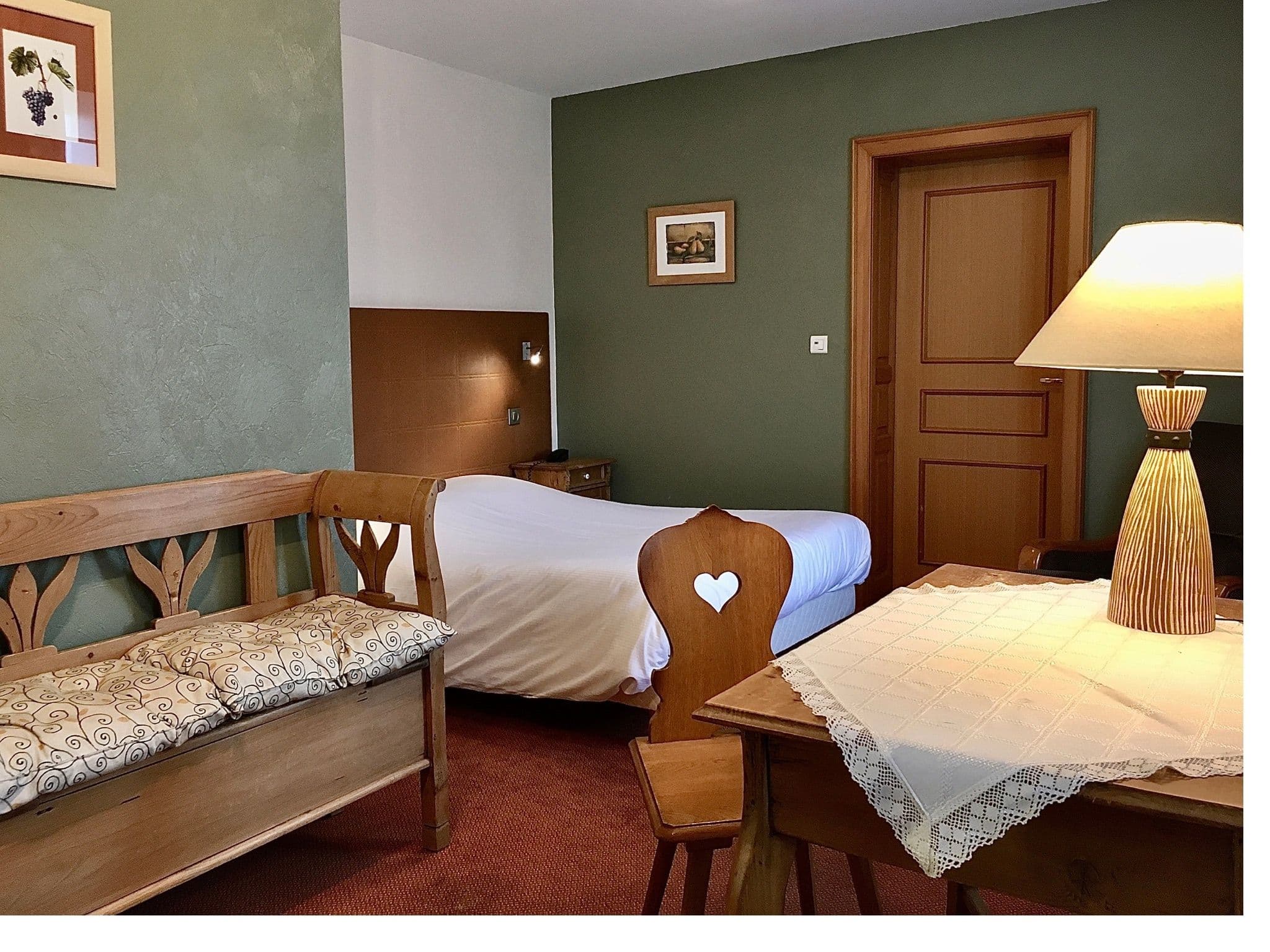 Chambre traditionnelle alsacienne avec mobilier en bois — banc sculpté avec coussins, chaise à dossier en forme de cœur, petite table avec nappe en dentelle et lampe, lit soigneusement fait contre un mur vert.