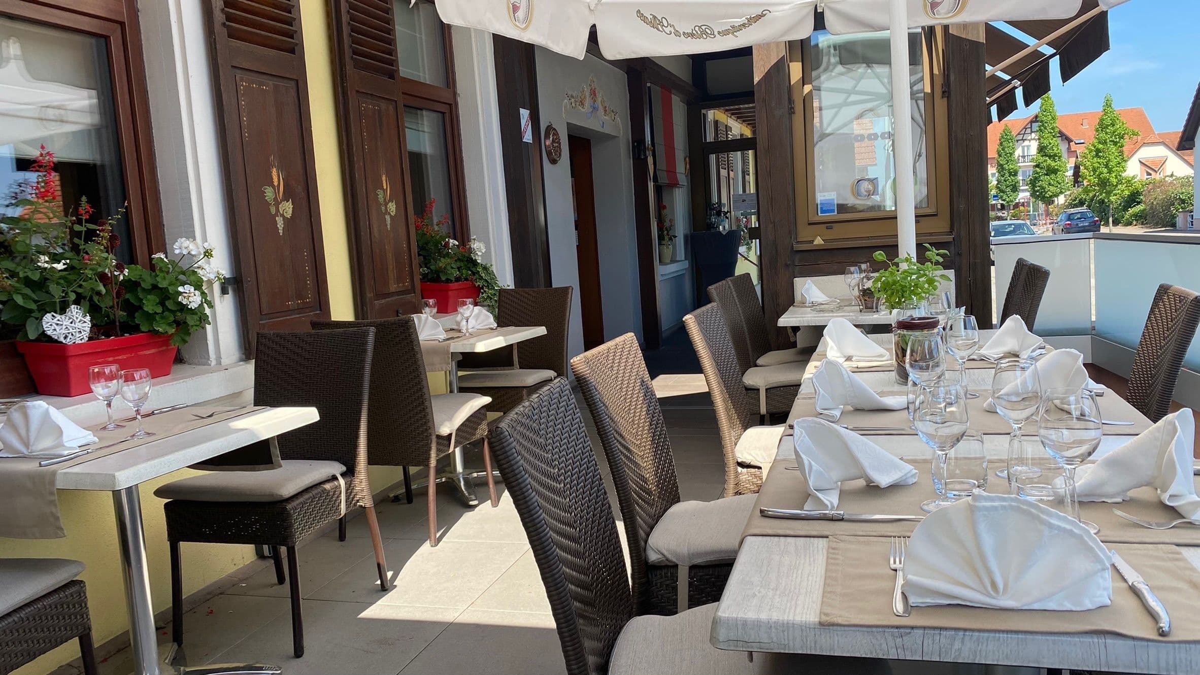 Terrasse ensoleillée du restaurant de l'hôtel avec tables dressées, serviettes blanches pliées et chaises en osier devant une façade à colombages et jardinières fleuries.