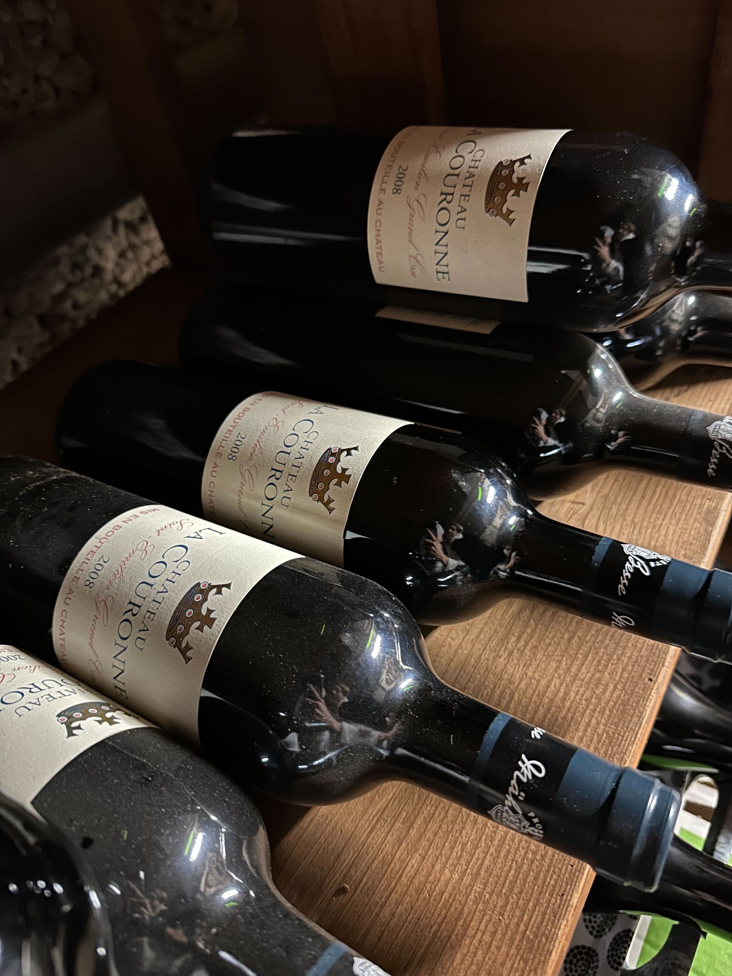 Gros plan sur plusieurs bouteilles de vin Château Couronne posées sur une clayette en bois, étiquettes vieillies et poussière laissant présager un stockage en cave.